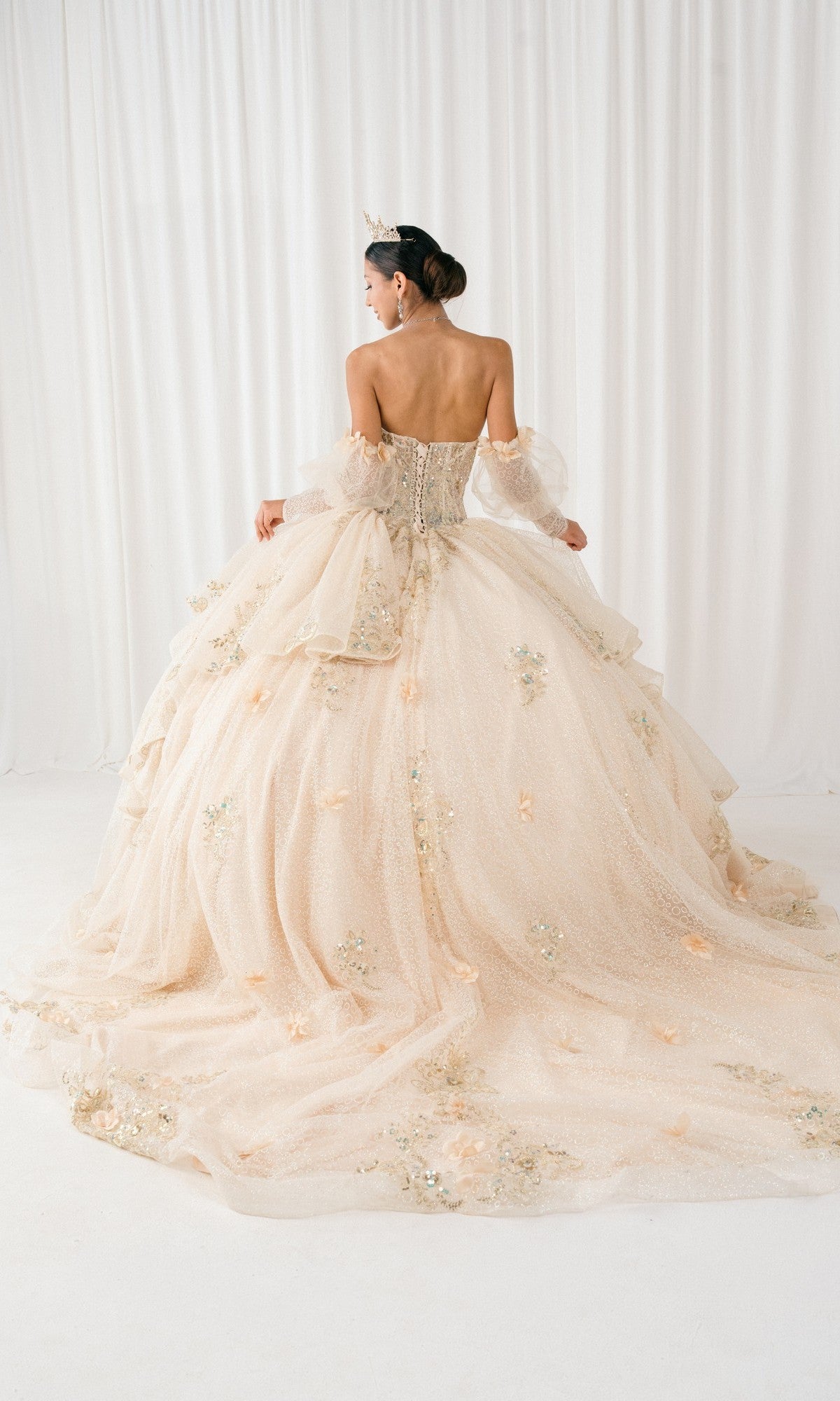 A woman wearing the Quinceanera Dress 8385J by Cinderella Couture, featuring a strapless embellished corset bodice and long detailed train, stands before a white curtain with a tiara adorning her head.